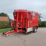 Large red farming trailer parked on a paved road beside hedges and houses under a cloudy sky, with hoses and equipment attached to its side.