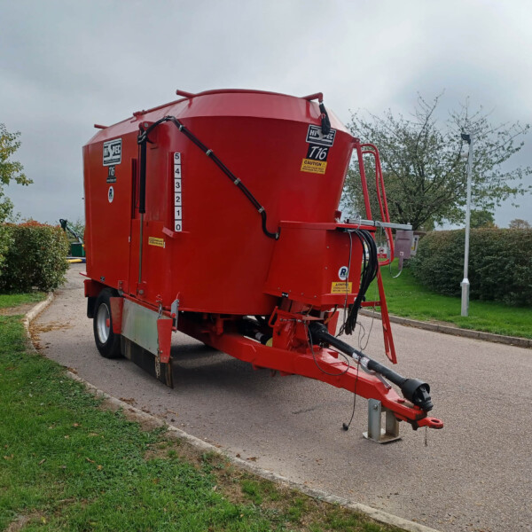 Large red agricultural trailer on wheels in a parking lot, with hoses and a hitch, under a cloudy sky.
