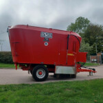 Red agricultural grain hopper/trailer on a truck in a parking lot with green grass and cloudy sky behind it