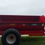 Red farm trailer with large tire, parked on grass with low buildings in the background.