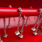 Red metal pipe with multiple chains and metal padlocks hanging along its length against a red background in an industrial scene.
