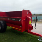 Red agricultural trailer with large black wheels parked on grass near a farm yard, showing its dump bed and hitch.