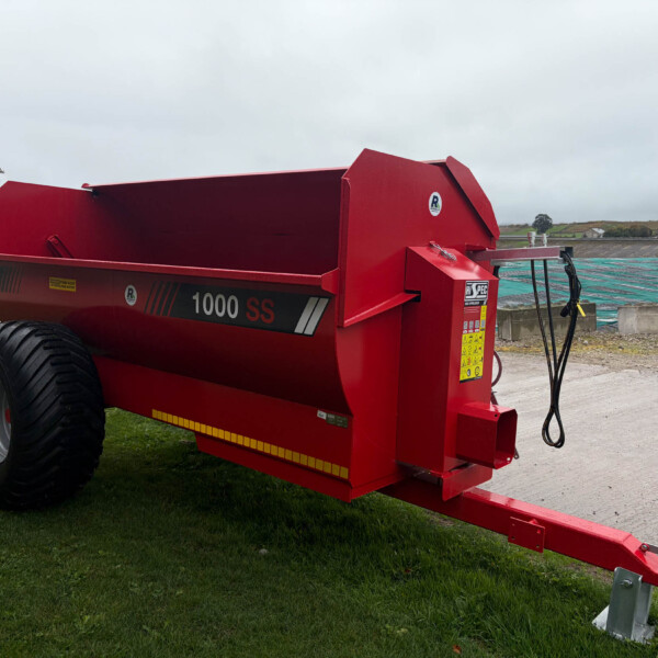 Red agricultural trailer with large black wheels parked on grass near a farm yard, showing its dump bed and hitch.