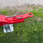 Close-up of a red metal towing or hitch mechanism with a curved silver latch resting on grass and damp soil nearby.