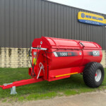 Red 1000 SS agricultural slurry tanker on a trailer, parked beside a dark corrugated building with a New Holland Agriculture sign above.
