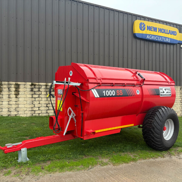 Red 1000 SS agricultural slurry tanker on a trailer, parked beside a dark corrugated building with a New Holland Agriculture sign above.
