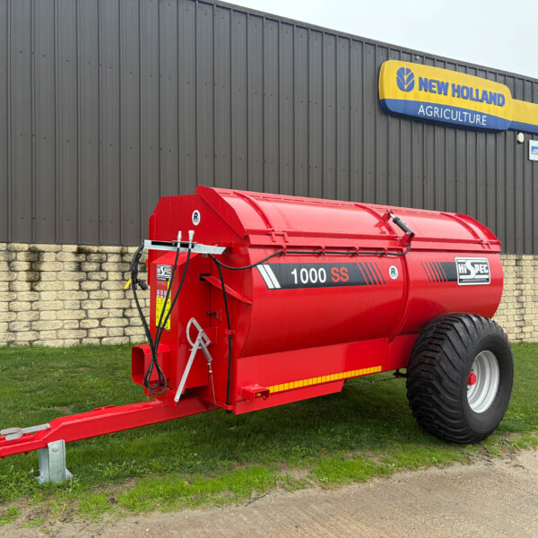 Red agricultural baler or forage harvester parked on grass beside a gray industrial wall with 'New Holland Agriculture' signage.
