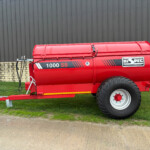 Red agricultural grain trailer with 1000 SS branding sits on a grassy area beside a brick wall and gray metal building, wheel visible at right edge.