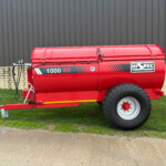 Red agricultural trailer with large rear wheel, parked on grass beside a stone wall and gray metal building.