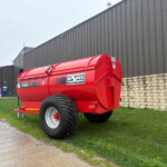 Red Hi-Spec 1000SS agricultural trailer with large tires parked on grass beside a gray metal warehouse and stone wall.