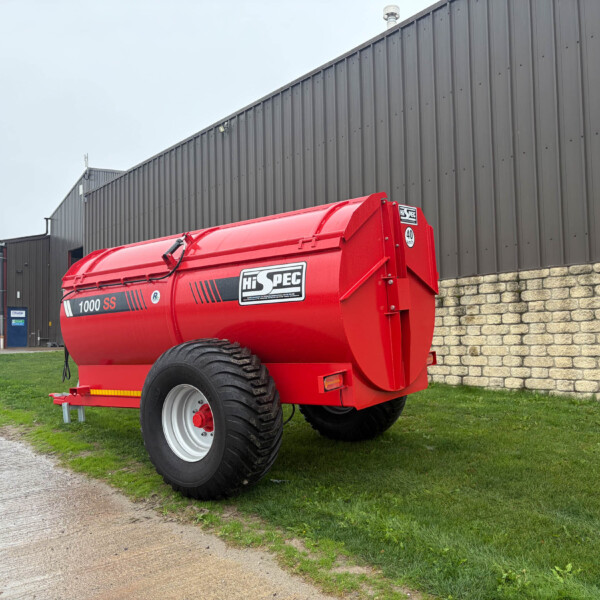 Red Hi-Spec 1000SS agricultural trailer with large tires parked on grass beside a gray metal warehouse and stone wall.