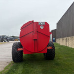 Rear view of a large red farming implement with 40 km/h label, parked beside a gray metal building on a gravel lot.