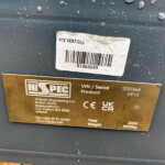 White barcode label reading 'V12 VERTICAL' on a dark metal surface, above a gold manufacturer plate on an industrial device.