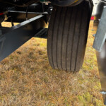 View under a heavy metal chassis with a large ribbed tire and steel frame, parked on dry grass.