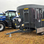 Line of blue New Holland tractors parked beside a large dark gray grain wagon on a dry grassy field