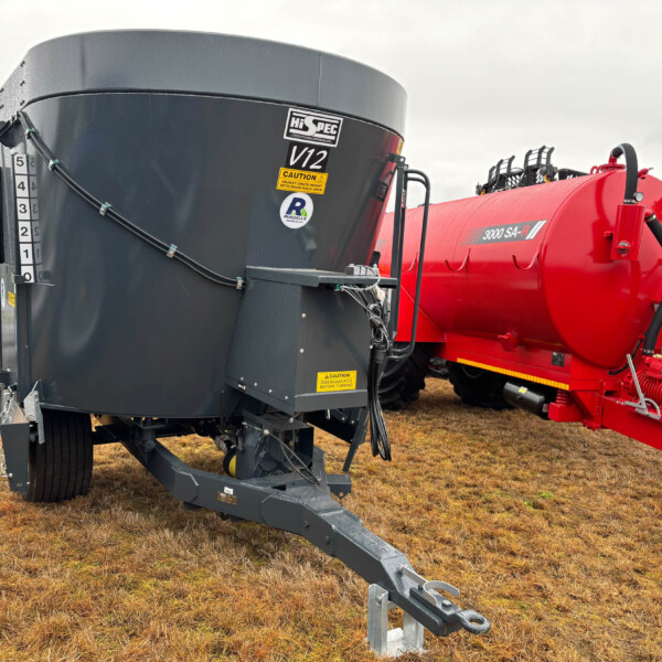 Large gray industrial mixer/hopper attached to a red tanker trailer in a field, with hoses and labels visible.