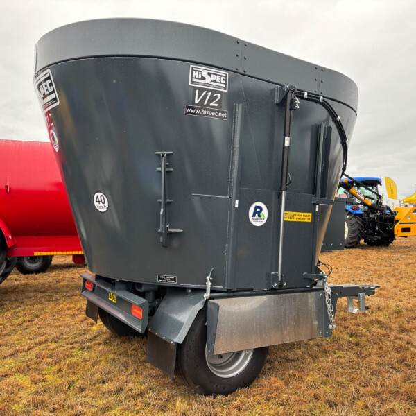 Close-up of a large gray agricultural trailer with 'V12' and Hispec branding, parked at an outdoor show with a red tractor nearby.
