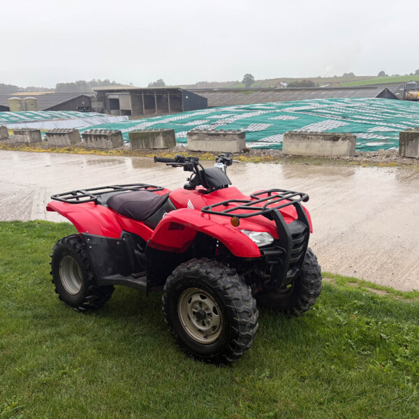 Red ATV parked on a wet concrete surface with green grass in the foreground and farm buildings in the background, including a large teal tarp-covered area behind.
