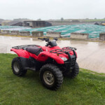 Red ATV parked on grass in a farmyard with sheds and green tarps in the background on a rainy day.