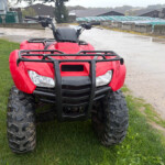 Front view of a red Honda ATV with a black front rack, parked on wet grass with a muddy industrial area behind it.
