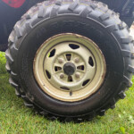 Close-up of a large off-road tire with deep tread on a weathered metal wheel, resting on grass.