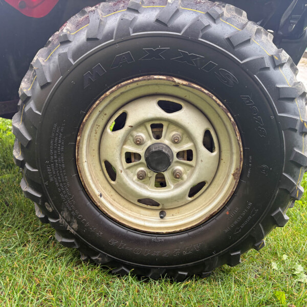 Close-up of a large off-road tire with deep tread on a weathered metal wheel, resting on grass.