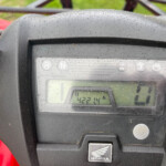 Close-up of a motorcycle instrument panel showing gear indicators (R, N) and a Honda emblem, with droplets on the casing.
