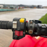 Close-up of a motorcycle handlebar with a yellow kill switch and other controls on a wet grip; blurred track in the background.