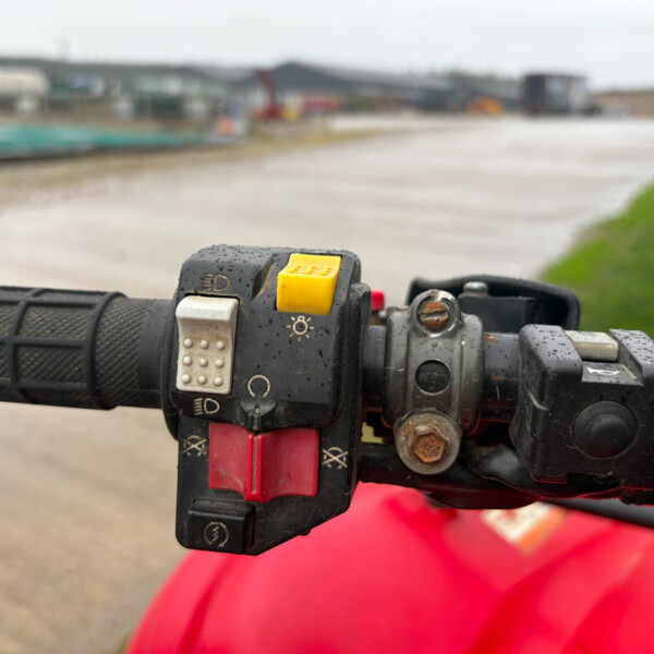 Close-up of a motorcycle handlebar with a yellow kill switch and other controls on a wet grip; blurred track in the background.