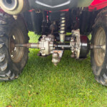 Underbelly view of a muddy tractor rear axle with suspension springs and a hitch, seen between large muddy tires on grass.