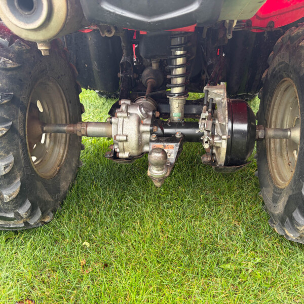 Underbelly view of a muddy tractor rear axle with suspension springs and a hitch, seen between large muddy tires on grass.