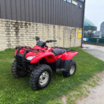 Red ATV parked on grassy area beside a stone wall and industrial building façade
