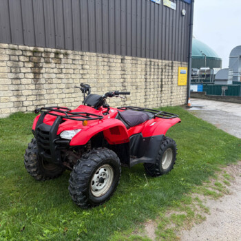 Red ATV parked on grass beside a stone wall and industrial metal building in the background.