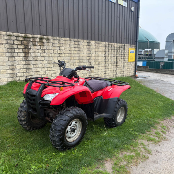 Red ATV parked on grass beside a stone wall and industrial metal building in the background.