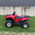 Red Honda ATV parked on a grassy area in front of a light stone wall and dark metal siding, handlebars visible.