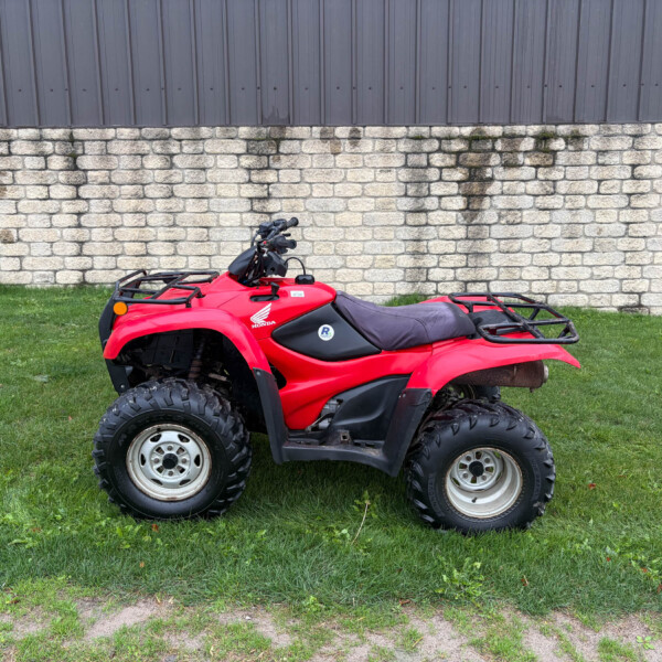Red Honda ATV parked on a grassy area in front of a light stone wall and dark metal siding, handlebars visible.