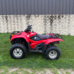 Red Honda ATV parked on grass in front of a stone wall and dark metal siding building behind it