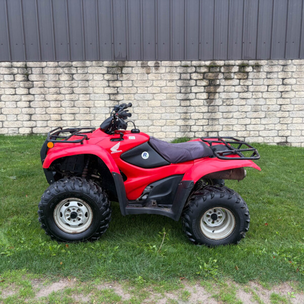 Red Honda ATV parked on grass in front of a stone wall and dark metal siding building behind it