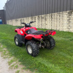 Red ATV parked on grass beside a beige stone wall and a dark metal warehouse building behind it.