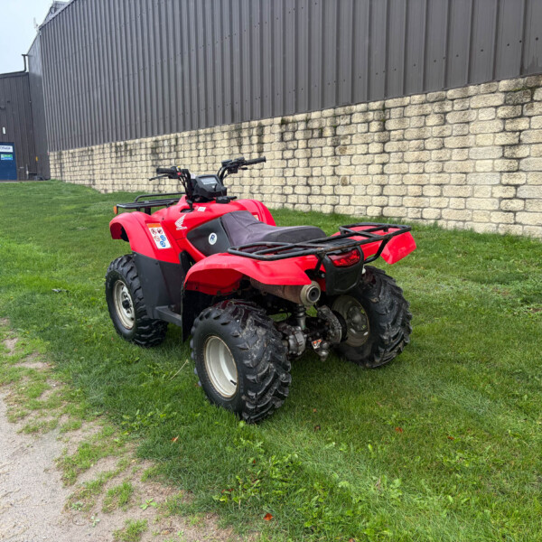 Red ATV parked on grass beside a beige stone wall and a dark metal warehouse building behind it.