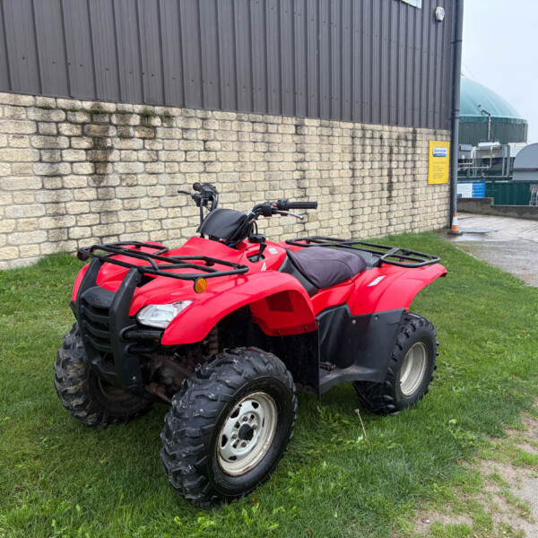 Two red ATVs parked on grass beside a stone wall with an industrial building in the background