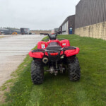 Red ATV parked on a grassy verge beside a concrete yard and large warehouse buildings in the background.