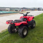 Red Honda ATV parked on a grassy verge beside a concrete area, with an industrial site and trucks in the distance behind it.