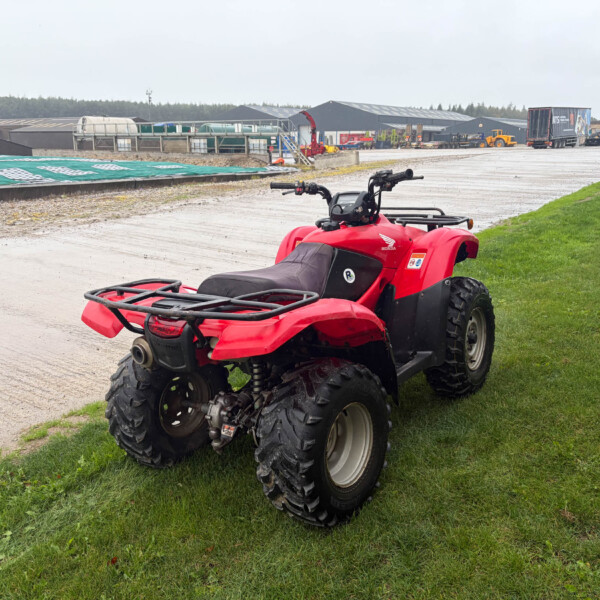 Red Honda ATV parked on a grassy verge beside a concrete area, with an industrial site and trucks in the distance behind it.