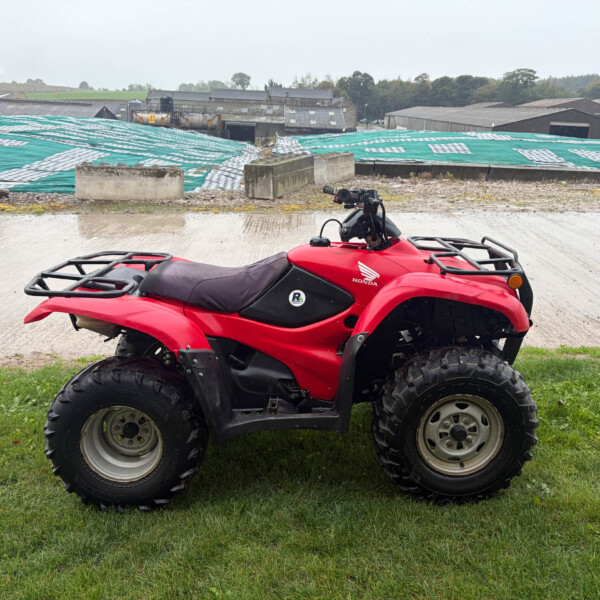 Red Honda ATV parked on a grassy area with industrial buildings and green tarps in the background.
