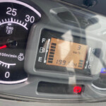 Close-up of a car dashboard: tachometer at 0, red needle resting near zero, and a digital display with warning indicators including the parking brake.
