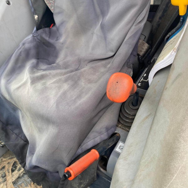 Interior of a construction machine cab with a dusty gray seat cover, orange joystick controls, and a foot pedal nearby