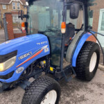 Blue Isuzu? Wait: actually a blue tractor (Iseki TG 6370) parked near a building, front and side views with large treaded tires and cabin doors visible.