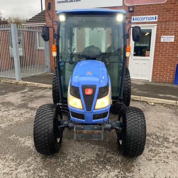 Front view of a blue tractor with headlights on, parked outside a brick building with Russell's sign and reception doors/signs in the background.