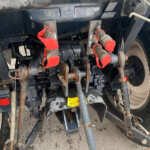 Rear view of a farming implement showing hydraulic hoses, red-handled coupling levers, and rusted metal framework on a gravel surface with a yellow warning sticker nearby